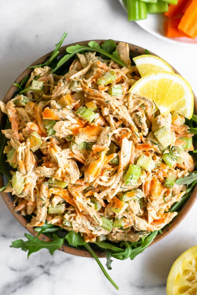 Healthy buffalo chicken salad in a bowl with arugula under it. It is garnished with lemon wedges and next to it is half a lemon and a plate with carrots and celery.