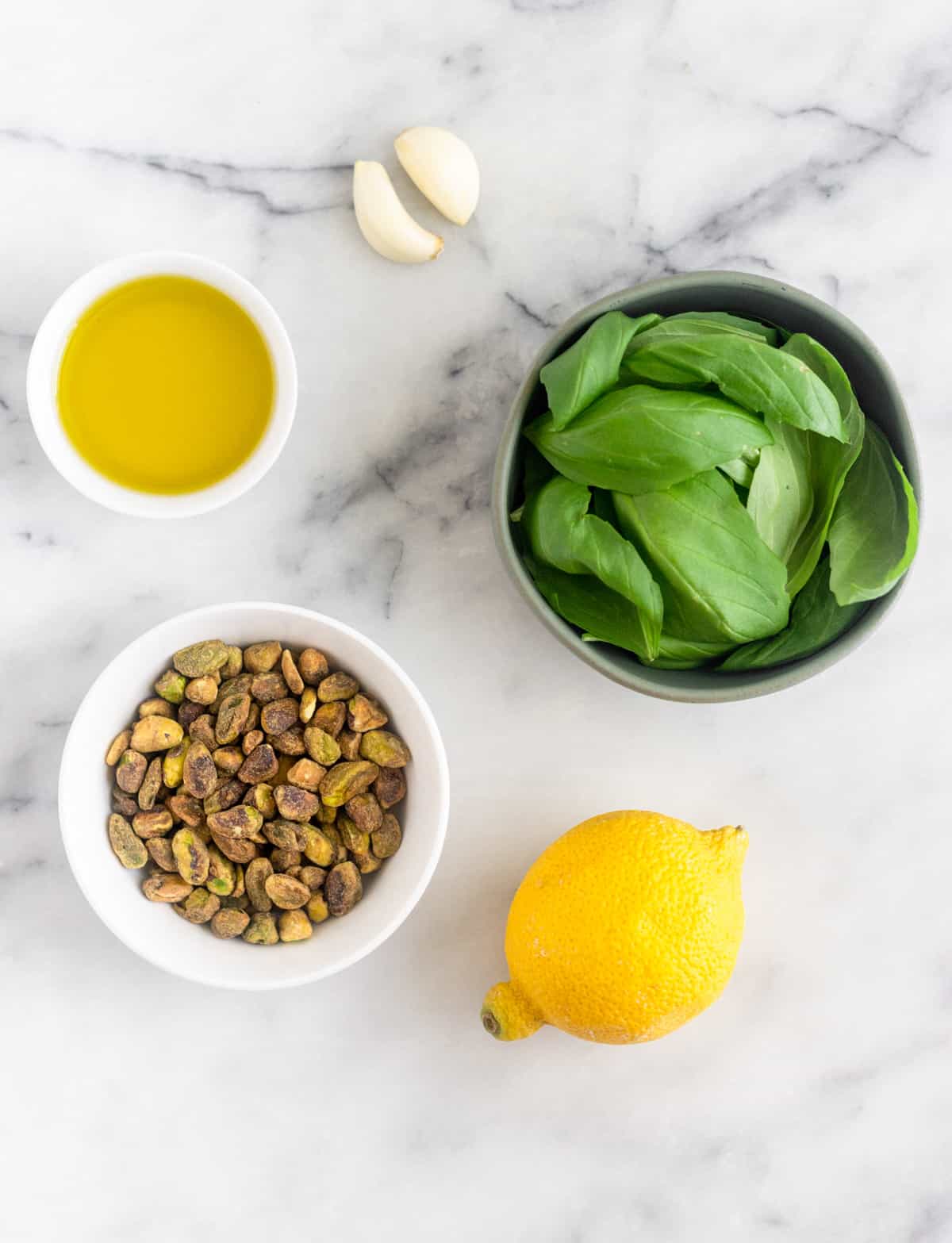 White counter with garlic, fresh basil in a bowl, a lemon, a bowl of shelled pistachios, and a bowl of olive oil on it