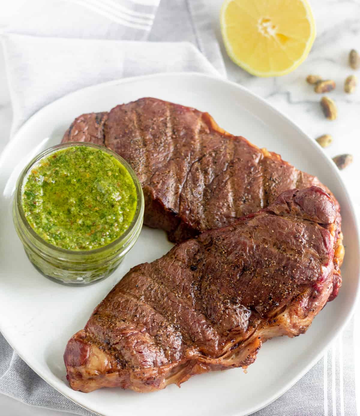 Two grilled steaks with a jar of pistachio pesto on a white plate with half a lemon and shelled pistachios behind it