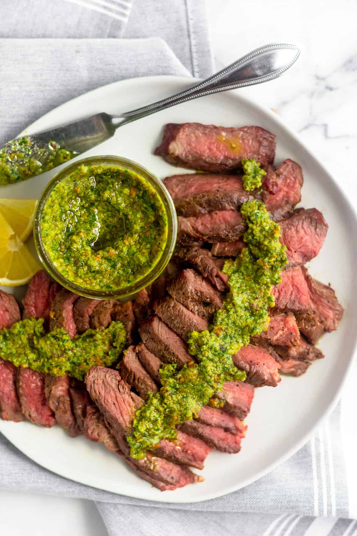 White plate with cut up grilled steak with pistachio pesto on top of it along with a jar of pistachio pesto and sliced lemon