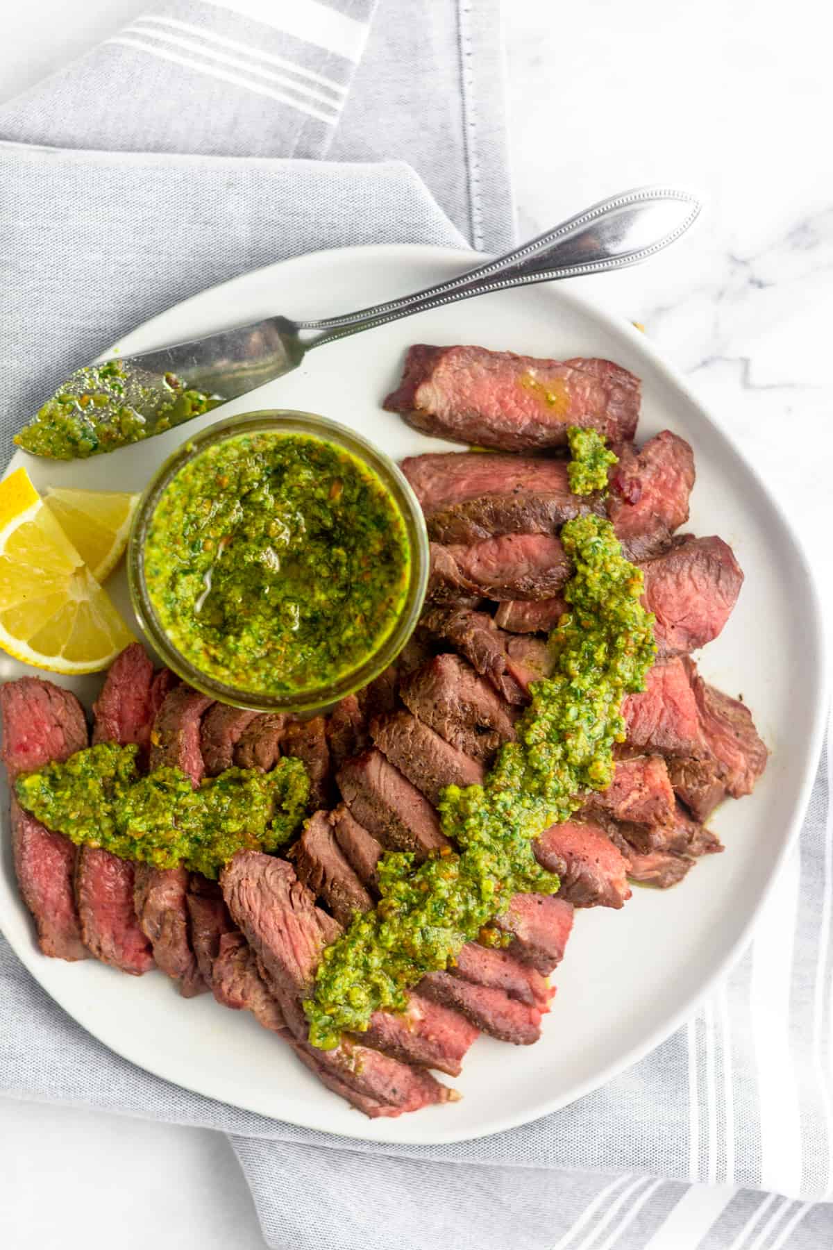 Overhead view of cut up grilled steak topped with pistachio pesto along with a jar of pesto, a small knife, and cut up lemons