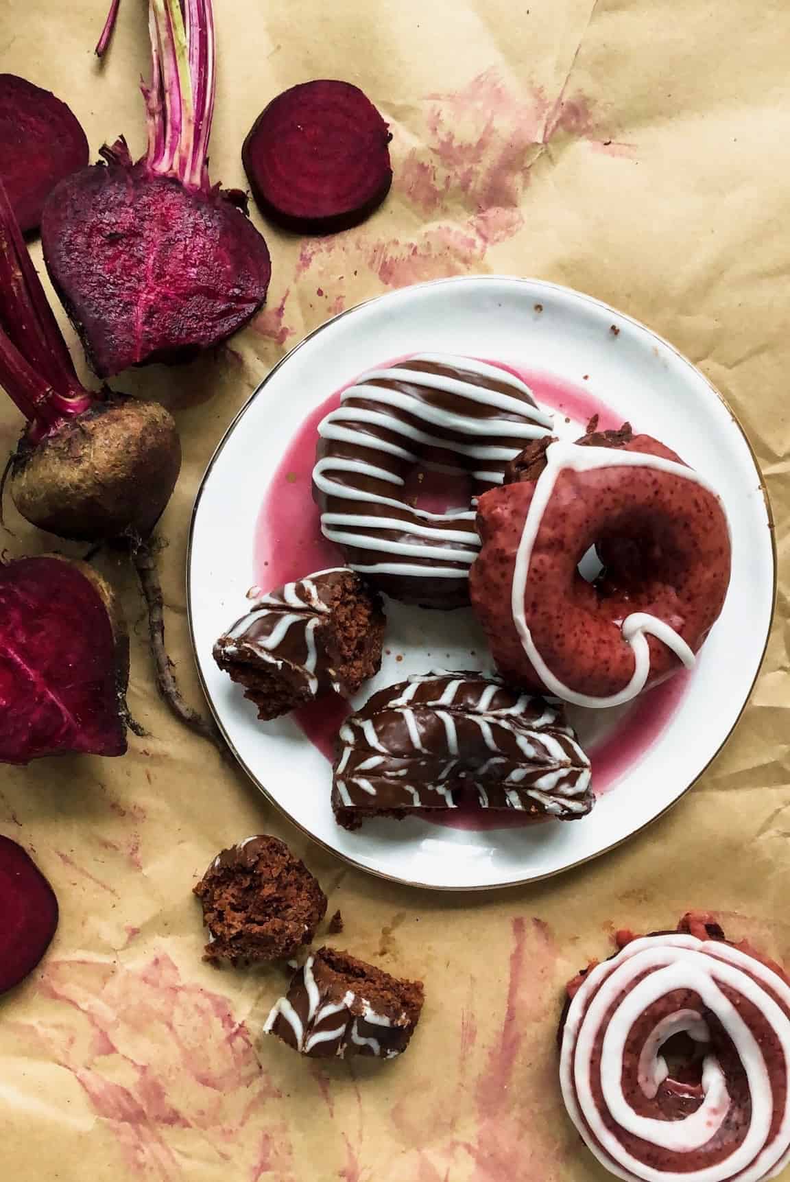 A white plate with 3 beet donuts on it, one cut open, surrounded by fresh raw beets on a brown paper.