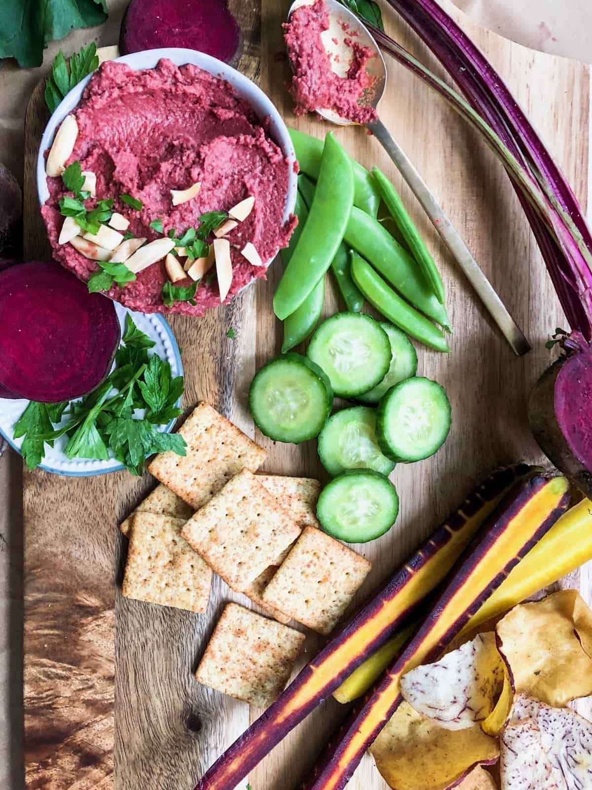 Spread of beet hummus, snap peas, cucumber spices, halved carrots, chips, and crackers on a wooden cutting board
