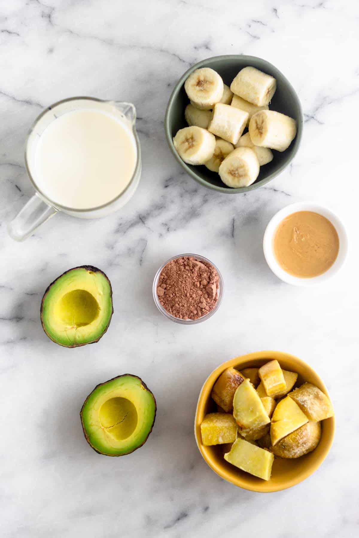 White marble slab with a cup of almond milk, frozen bananas, peanut butter, cacao powder, frozen sweet potatoes, and an avocado for a peanut butter frosty