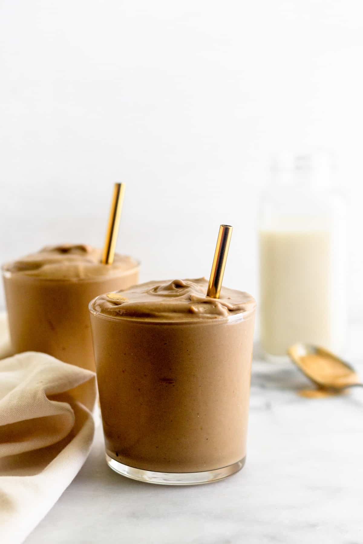 Straight on shot of two glass with peanut butter frosty in the with straws, a tan towel, glass of almond milk, and a spoon of peanut butter.