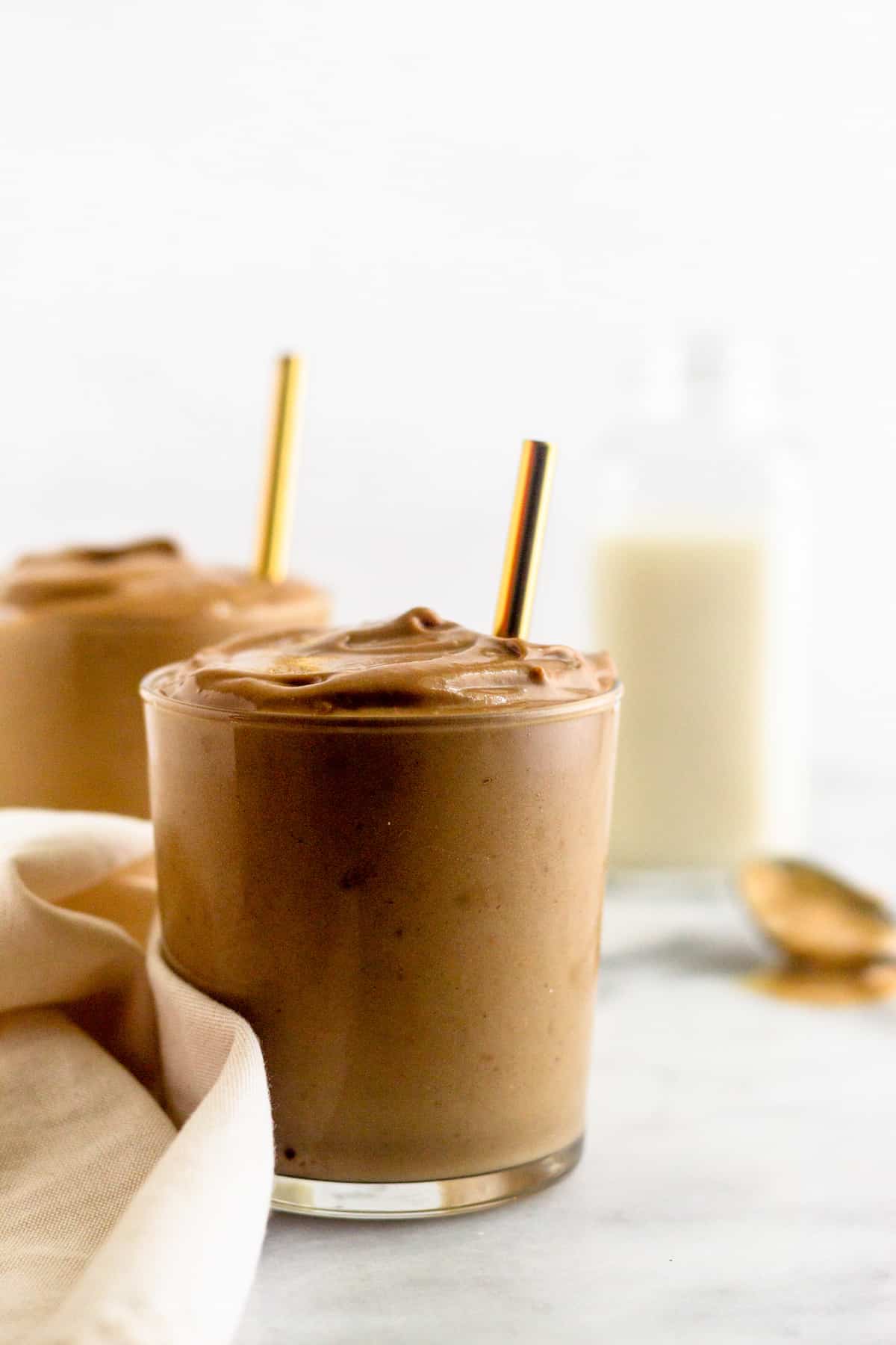 Two glass of a healthy dairy free peanut butter frosty with straws in them next to a white light brown towel. Behind them is a glass of almond milk and a spoon of peanut butter