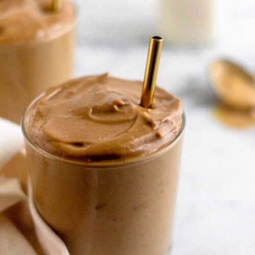 Glass filled with a healthy dairy free peanut butter frosty with a gold straw in it. Behind it is another glass with a frosty in it, a jar of almond milk, and a spoon of peanut butter.