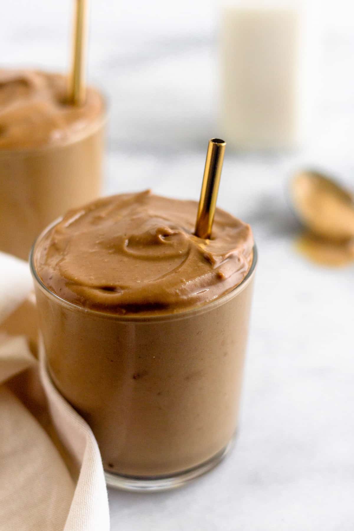 Glass filled with a healthy dairy free peanut butter frosty with a gold straw in it. Behind it is another glass with a frosty in it, a jar of almond milk, and a spoon of peanut butter.