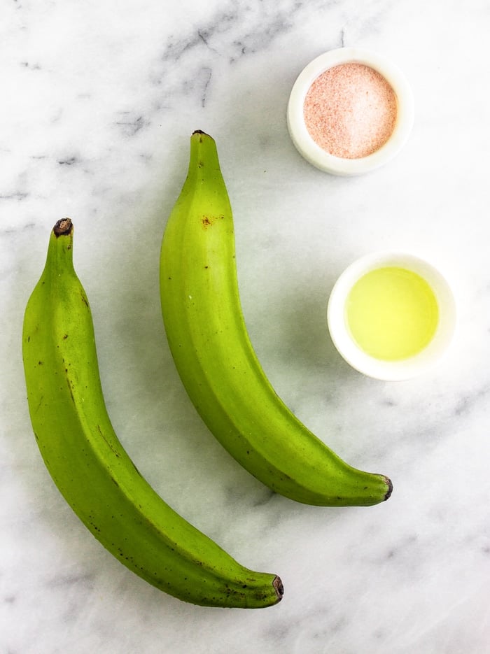 Overhead shot of two green plantains, a dish of salt, and a small dish of oil.