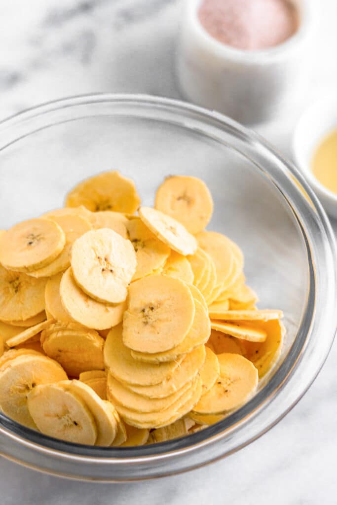 Sliced plantains in a large bowl. Behind them are a small bowl of oil and a small container of salt.