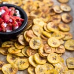 A large baking sheet with homemade baked plantain chips spread out all over with a small dish of salsa.