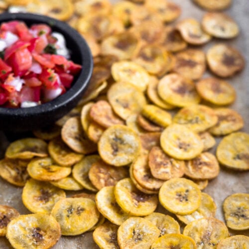 A large baking sheet with homemade baked plantain chips spread out all over with a small dish of salsa.