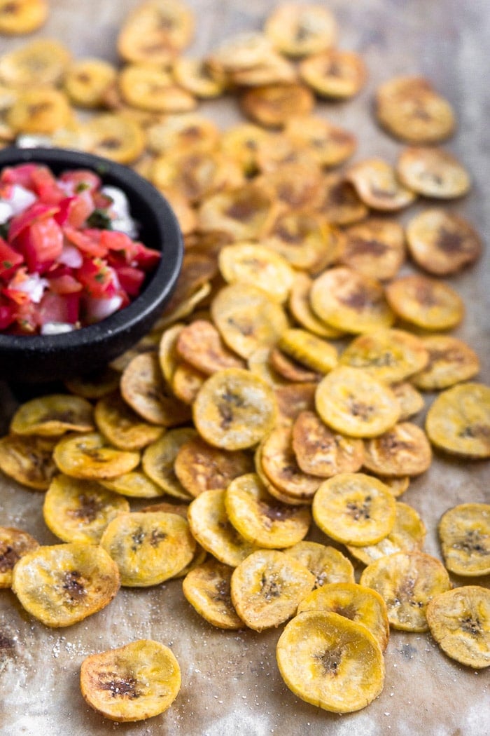 A large baking sheet with homemade baked plantain chips spread out all over with a small dish of salsa.