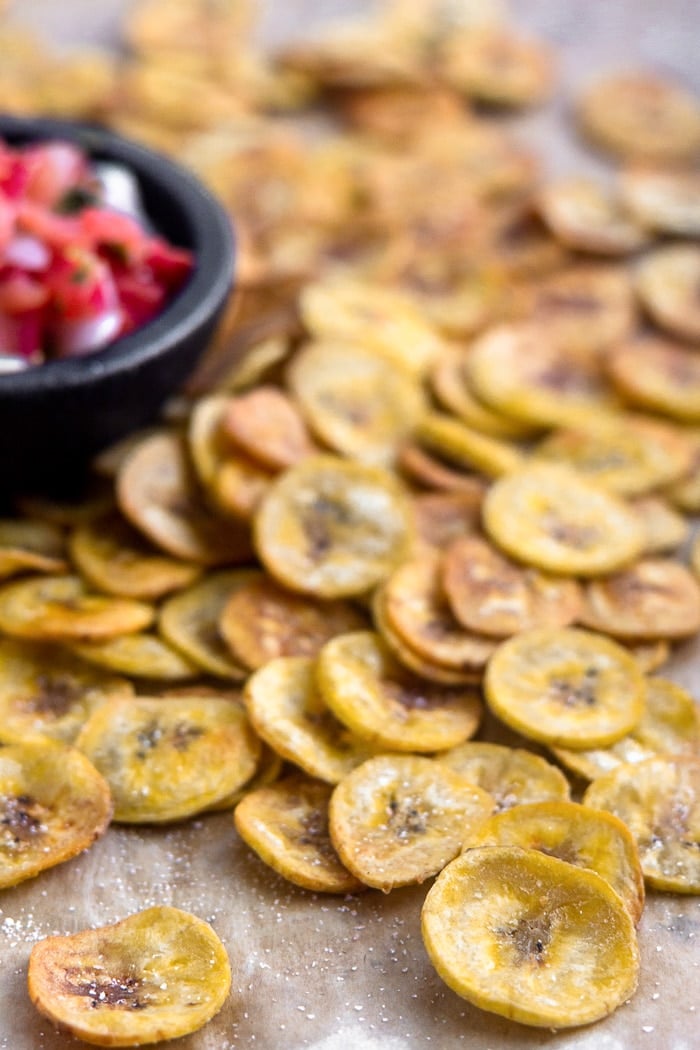 Homemade plantain chips spread out on a baking sheet with a bowl of salsa next to them.