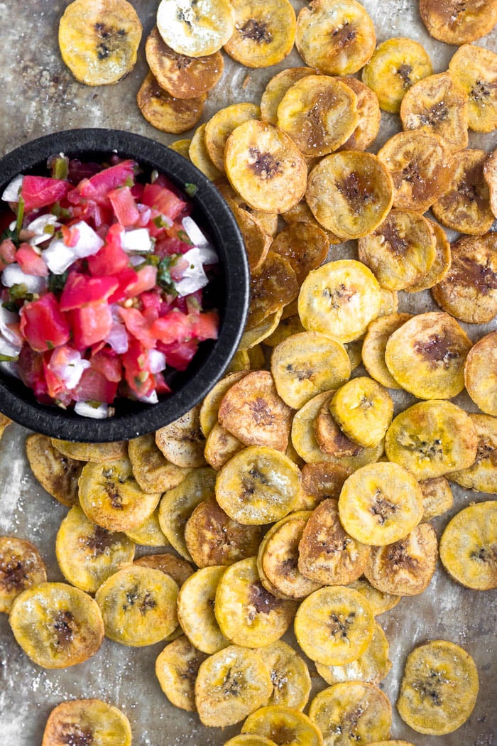 Overhead shot of baked plantain chips spread out on a baking sheet with a small dish of salsa.