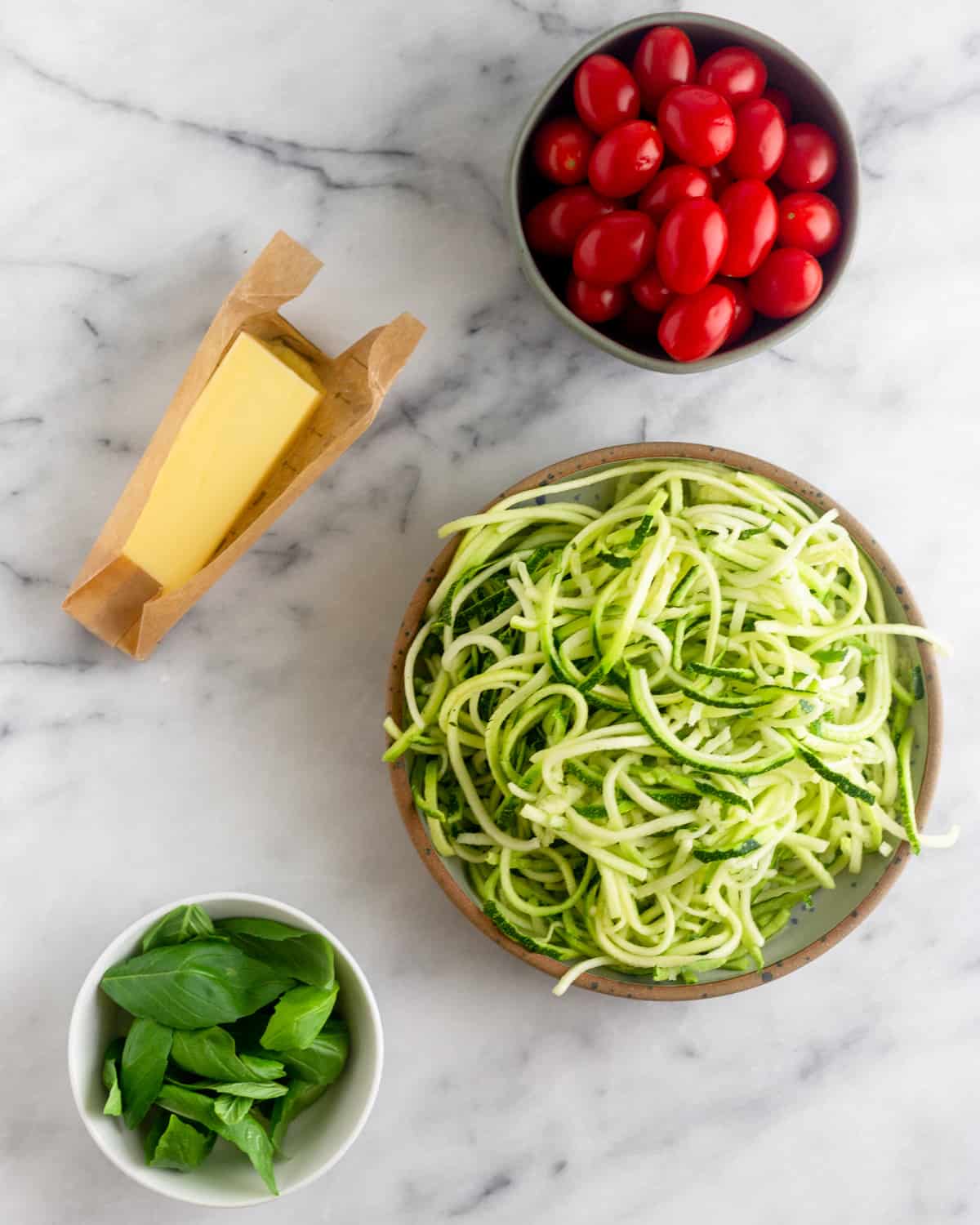 Bowl of cherry tomatoes, bowl of zucchini noodles, bowl of fresh basil, and a stick of butter on a white counter top