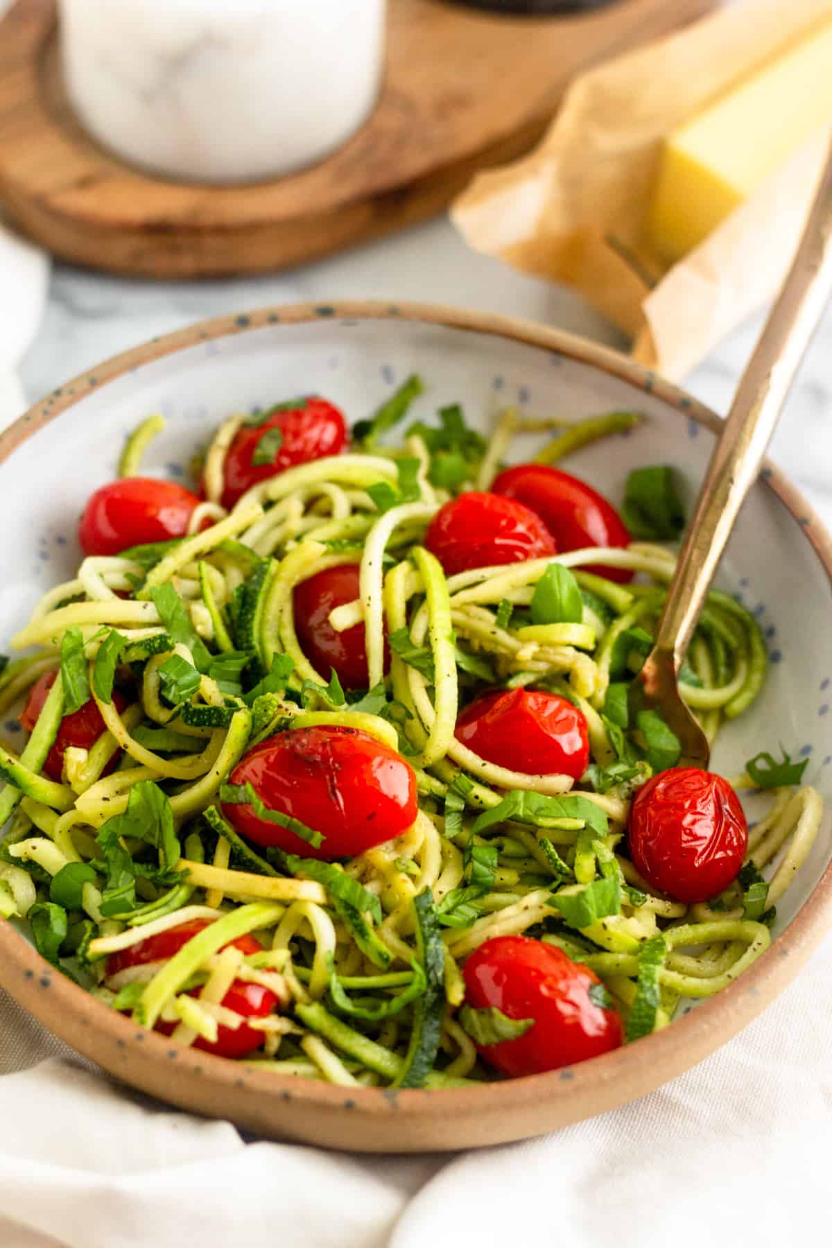 Bowl of zucchini noodles with blistered tomatoes and basil with a gold fork in it in front of a stick of butter and a salt holder