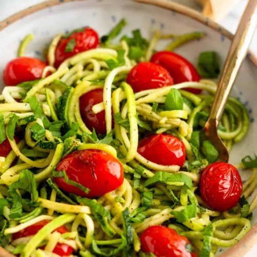 Bowl of zucchini noodles with blistered tomatoes and basil with a fork in it and a stick of butter behind it