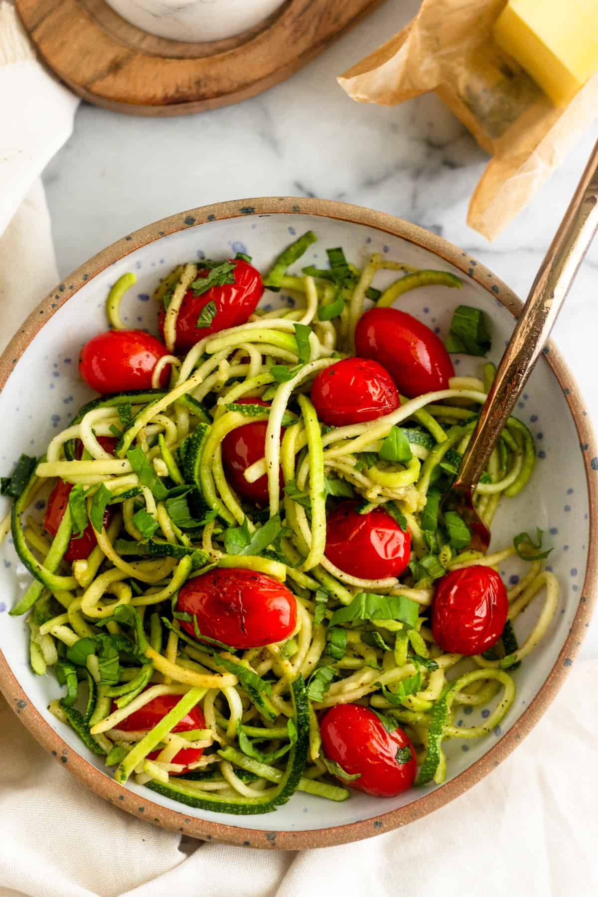 Overhead shot of a bowl of zucchini noodles with blistered tomatoes and basil with a gold fork in it next to a stick of butter and a salt holder