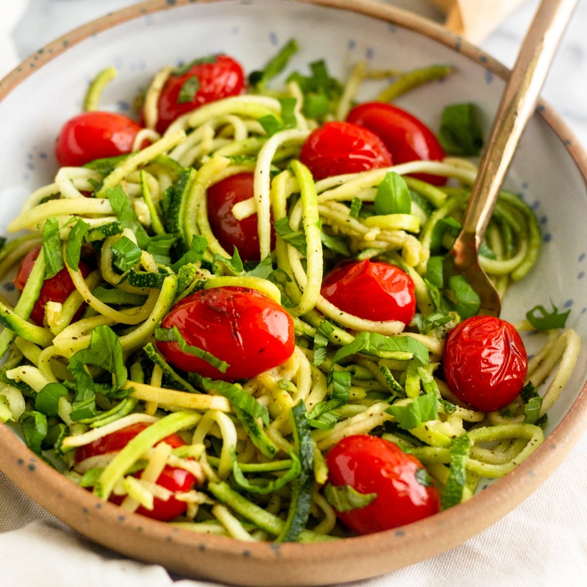 Bowl of zucchini noodles with blistered tomatoes and basil with a gold fork in it in front of a stick of butter and a salt holder