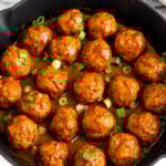 A large cast iron skillet filled with BBQ chicken meatballs covered in BBQ sauce and topped with green onions. Around it is some fresh parsley and a bowl of chopped green onions.