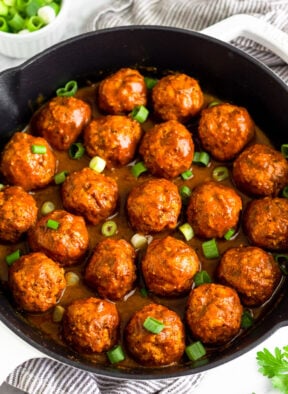 A large cast iron skillet filled with BBQ chicken meatballs covered in BBQ sauce and topped with green onions. Around it is some fresh parsley and a bowl of chopped green onions.