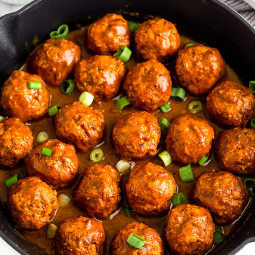 A large cast iron skillet filled with BBQ chicken meatballs covered in BBQ sauce and topped with green onions. Around it is some fresh parsley and a bowl of chopped green onions.