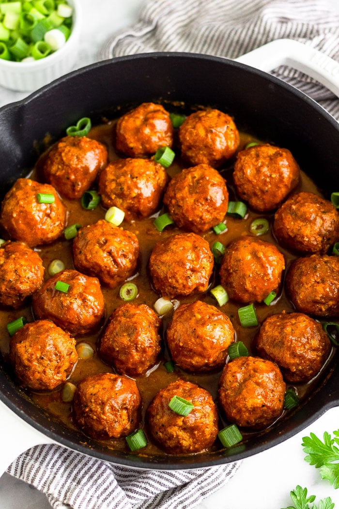 A large cast iron skillet filled with BBQ chicken meatballs covered in BBQ sauce and topped with green onions. Around it is some fresh parsley and a bowl of chopped green onions.
