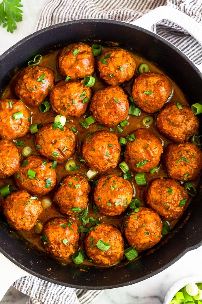 Overhead shot of BBQ chicken meatballs made in the instant pot. They are in a large cast iron with bbq sauce. chopped green onions, and chopped parsley. The pan is sitting on a towel with parsley and a bowl of green onions around it.