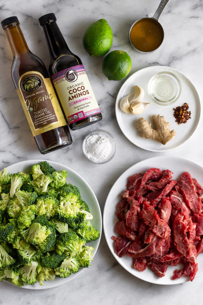 Overhead shot of a white counter with a bottle of sesame oil, bottle of coconut aminos, limes, small cup of broth, plate of garlic, ginger, spices, and a small bowl of oil, plate of thinly sliced steak, plate of broccoli florets, and a small bowl of flour.