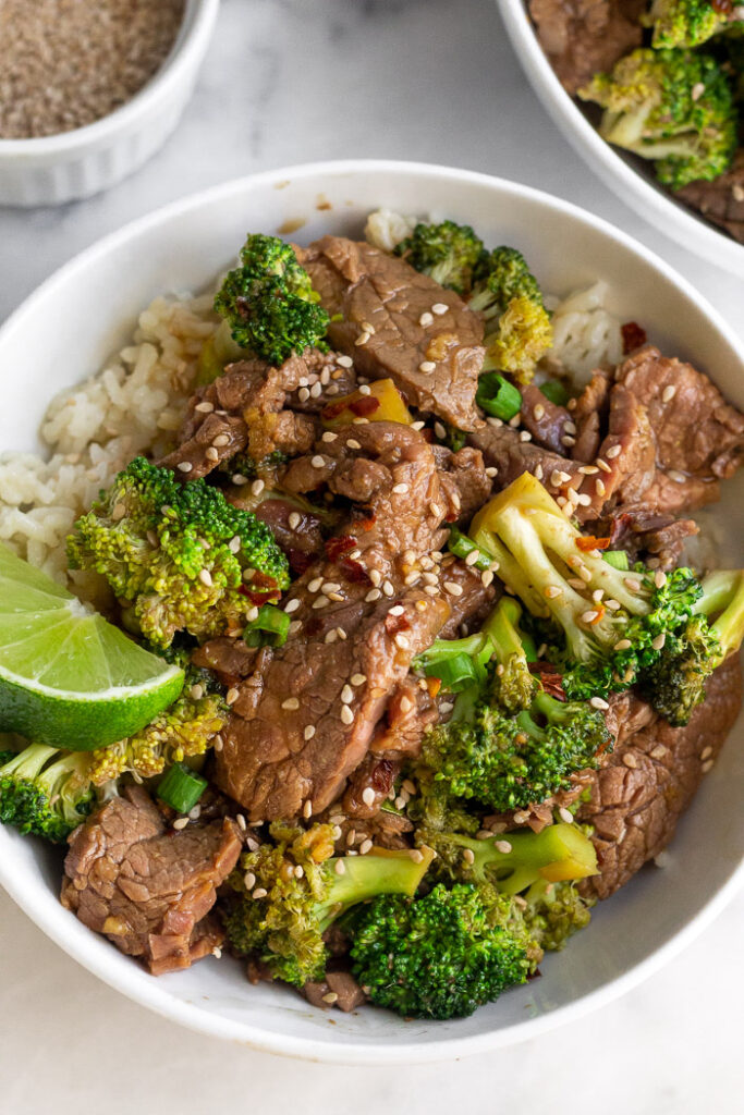 Beef and broccoli recipe in bowl on top of white rice. It is topped with sesame seeds, green onion, and a lime wedge. Behind it is another bowl and a small bowl of sesame seeds.