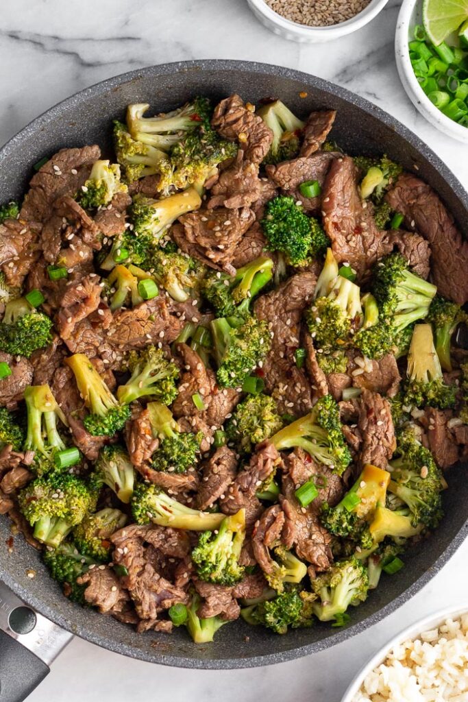 Easy beef and broccoli recipe in a large skillet. It is topped with sesame seeds. Next to the skillet is a small bowl of sesame seeds, a bowl green onion and limes, and a bowl of rice.