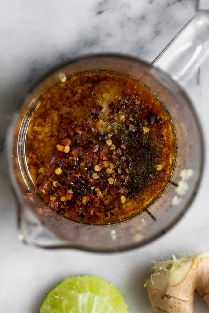 Measuring cup filled with coconut aminos, broth, lime juice, sesame oil, fresh ginger, fresh garlic, and red pepper flakes. Next to is in fresh ginger and half a lime.