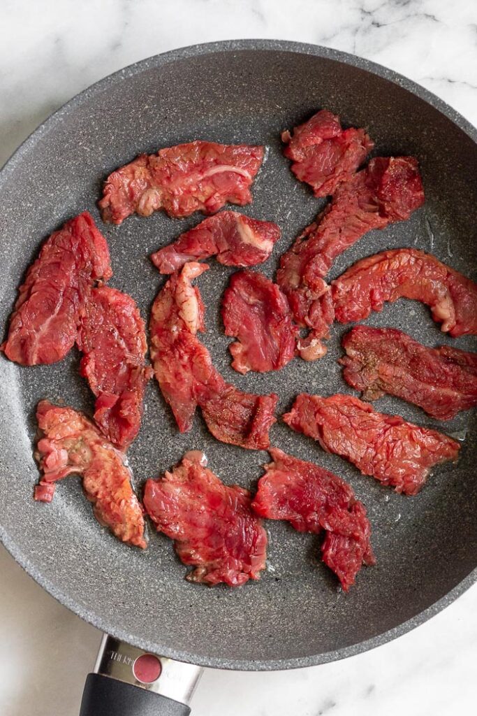 Large skillet with raw thinly sliced flank steak in it.