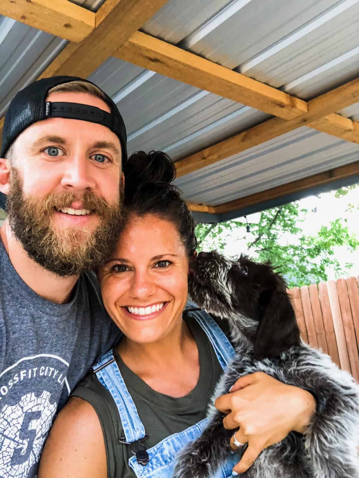 A girl, her fiancé, and their new puppy, who is licking her face, in a family photo outside