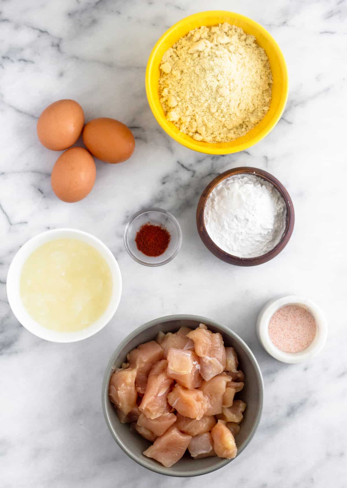 3 eggs, bowl of almond flour, bowl of tapioca flour, dish of salt, bowl of raw cubed chicken, bowl of coconut oil, and a bowl of paprika on a white counter.
