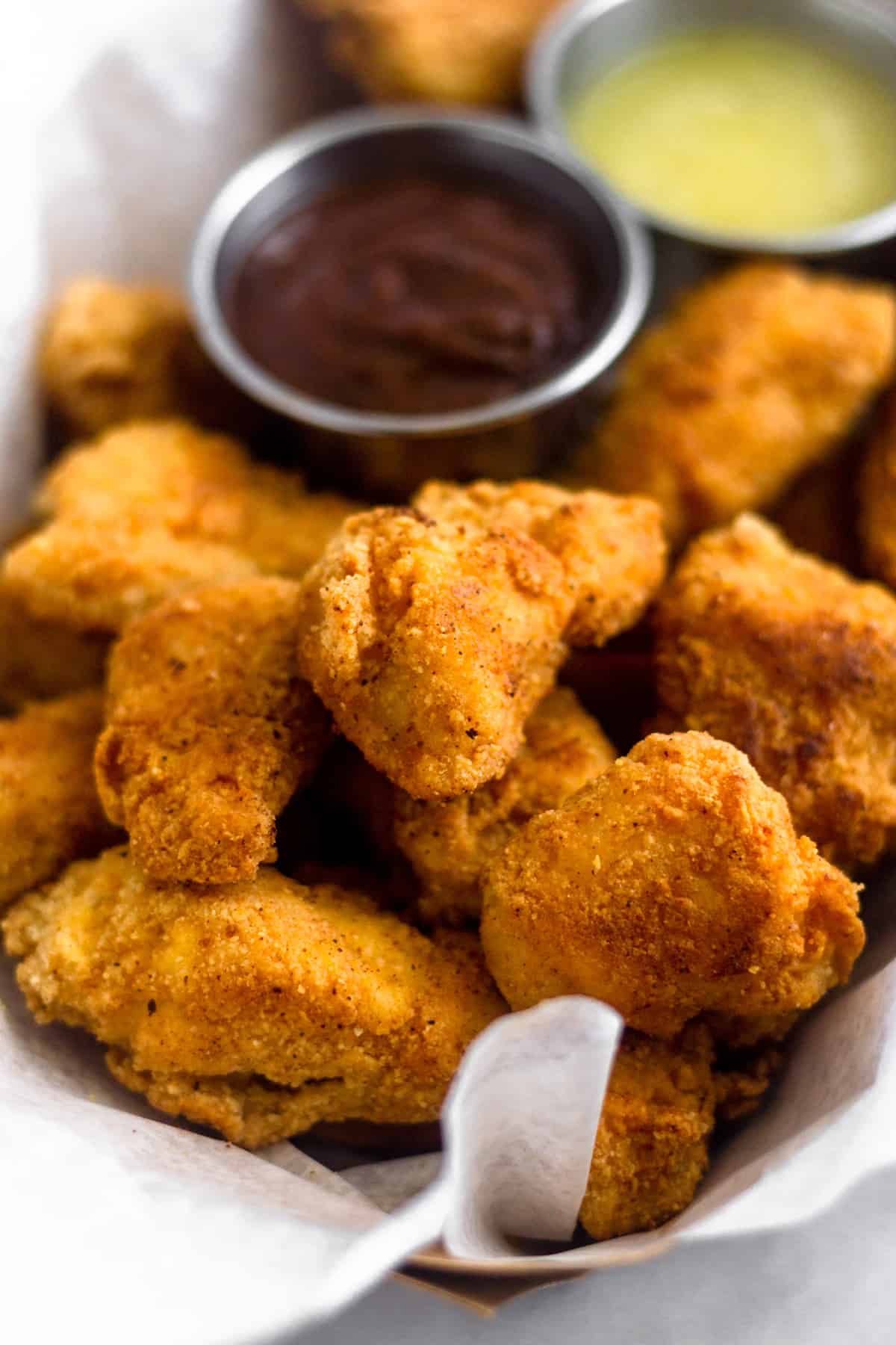 Close up of a basket of a bunch of whole30 and paleo popcorn chicken with dipping sauces behind it