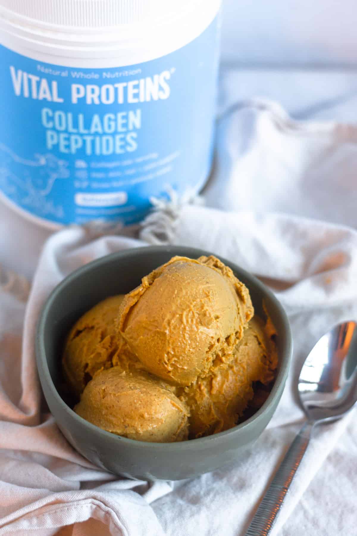 Bowl of paleo pumpkin pice cream on a tan linen, next to a spoon and a container of collagen behind it