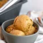 Bowl of dairy free and paleo pumpkin ice cream on a tan linen with a pan of pumpkin ice cream and a spoon behind it.