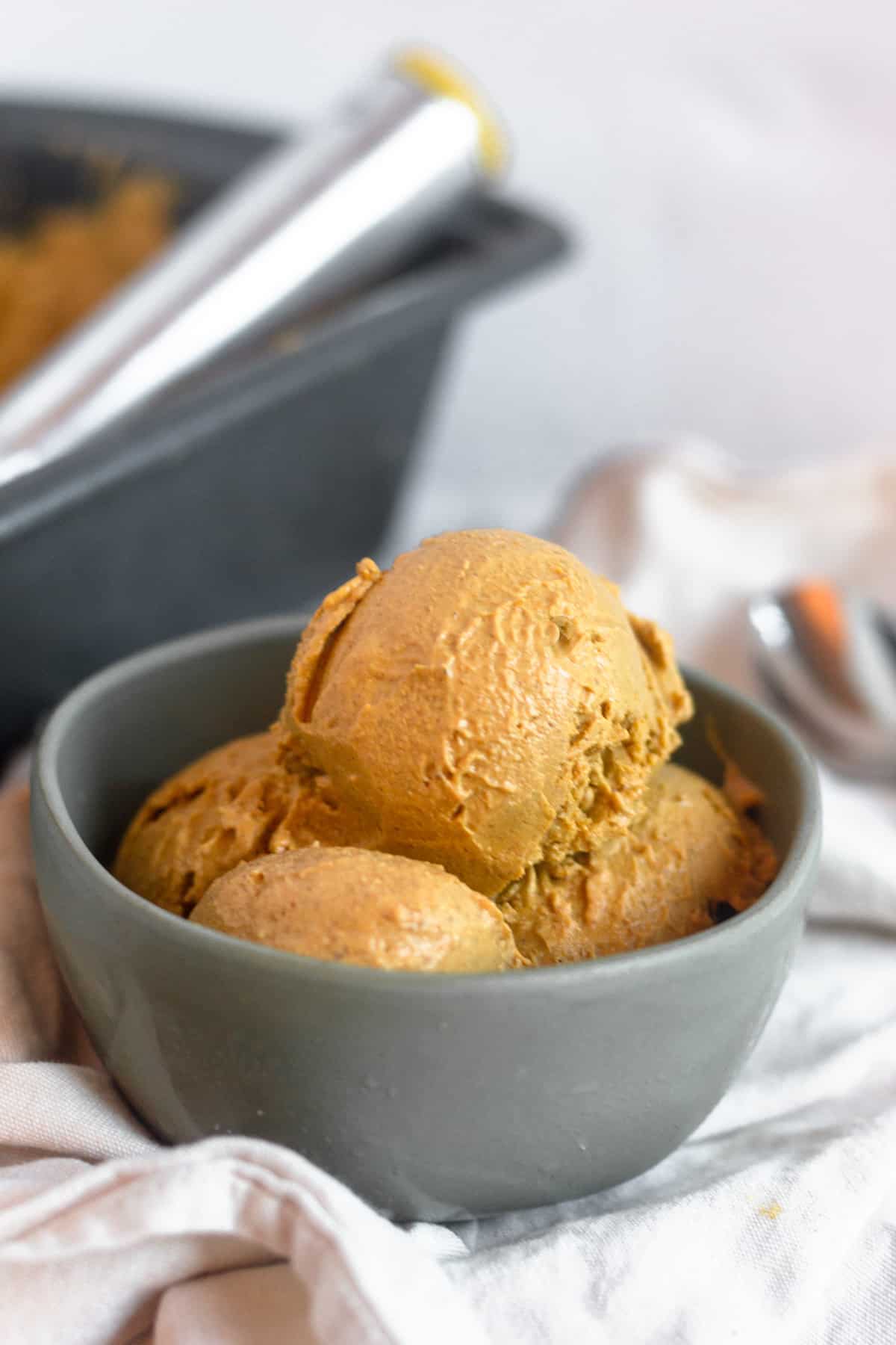 Bowl of dairy free and paleo pumpkin ice cream on a tan linen with a pan of pumpkin ice cream and a spoon behind it.