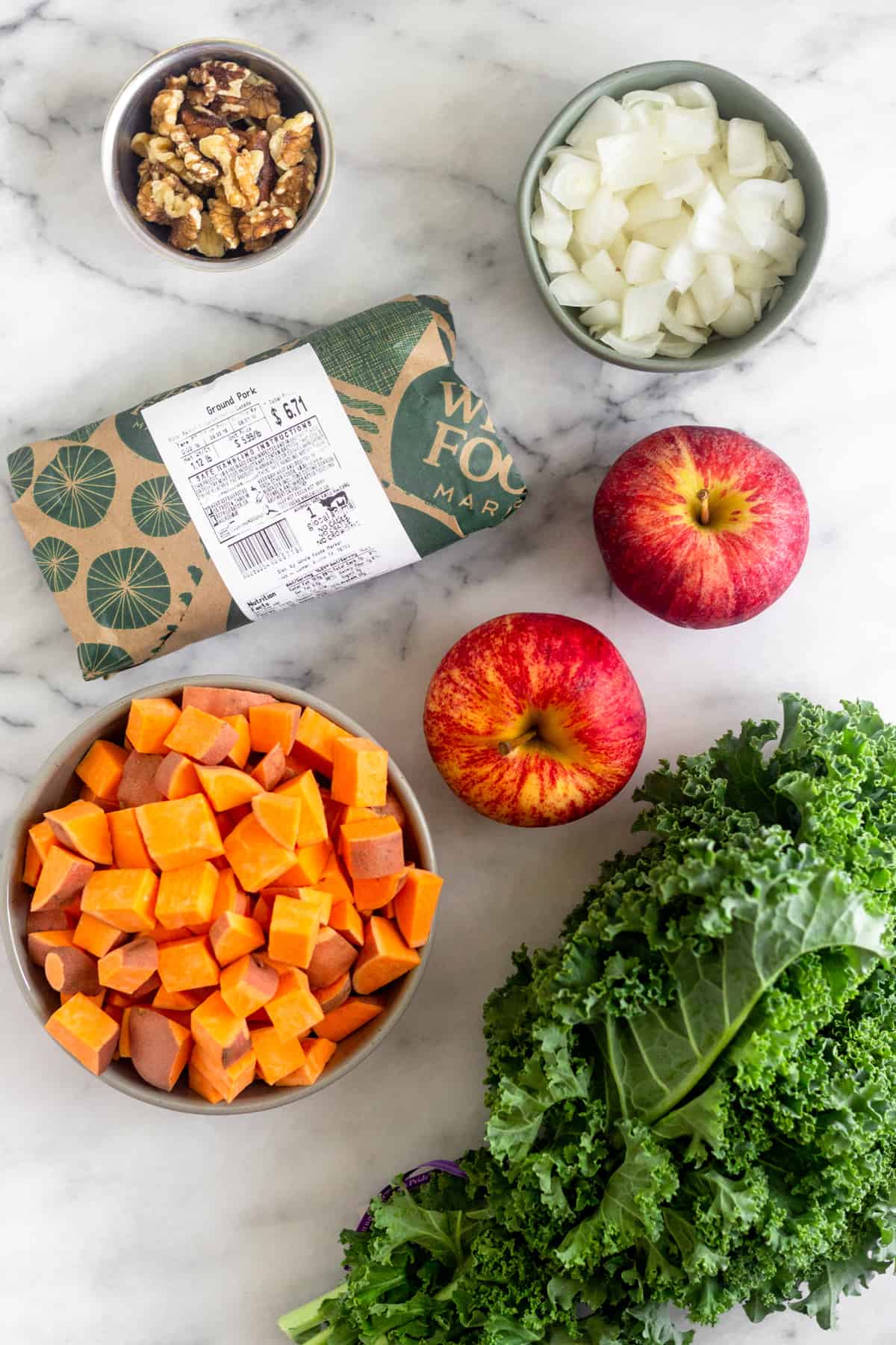 White counter with a small bowl of walnuts, bowl of diced onions, 2 apples, bunch of kale, bowl of diced sweet potatoes, and a package of ground pork.
