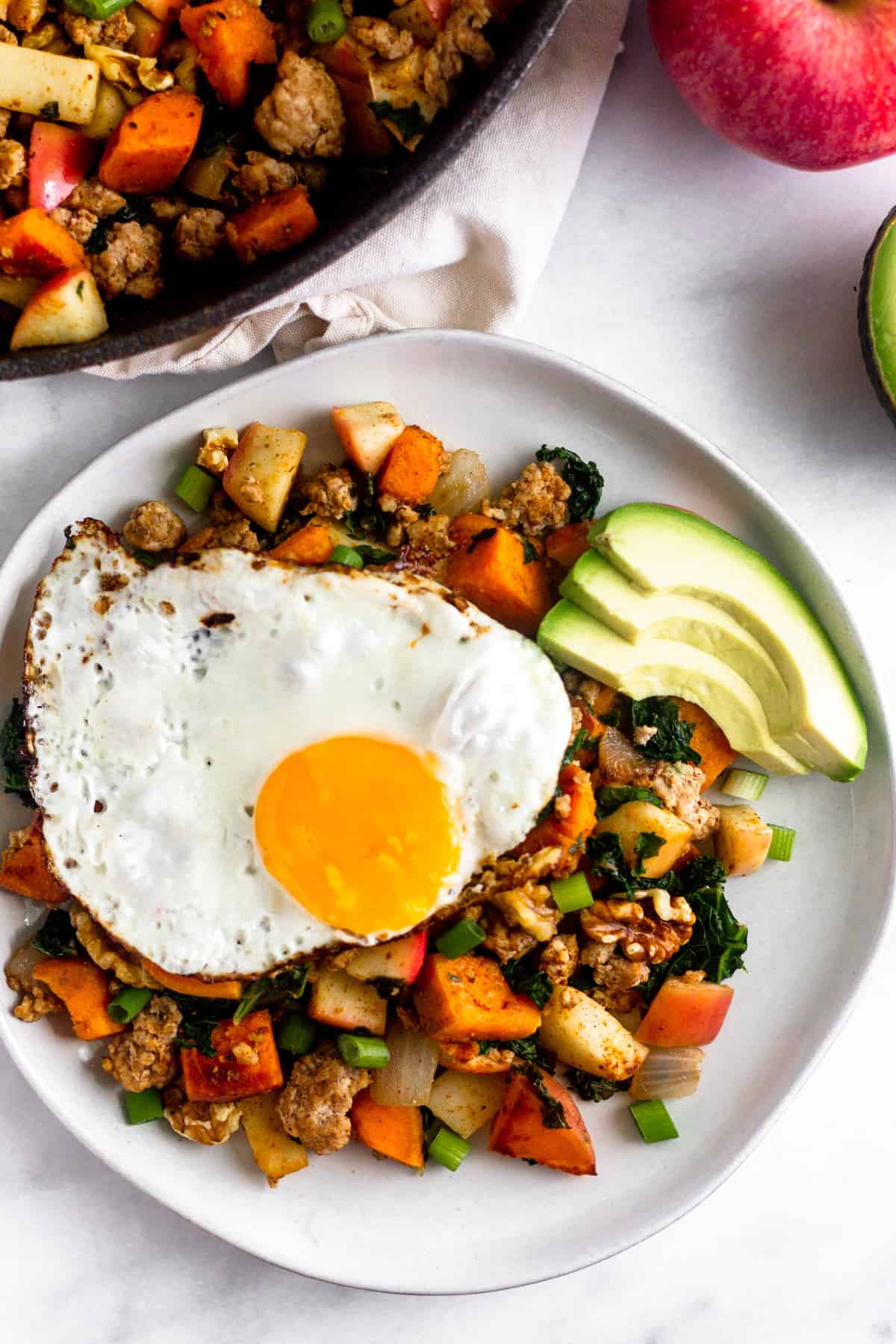 Overhead shot of a white plate filled with paleo sweet potato hash with sausage and apples topped with a fried egg and avocado. Next to it is a large pan of the hash, an apple, and half an avocado.