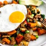 White plate on a tan linen with sweet potato sausage hash topped with a fried egg and sliced avocado.