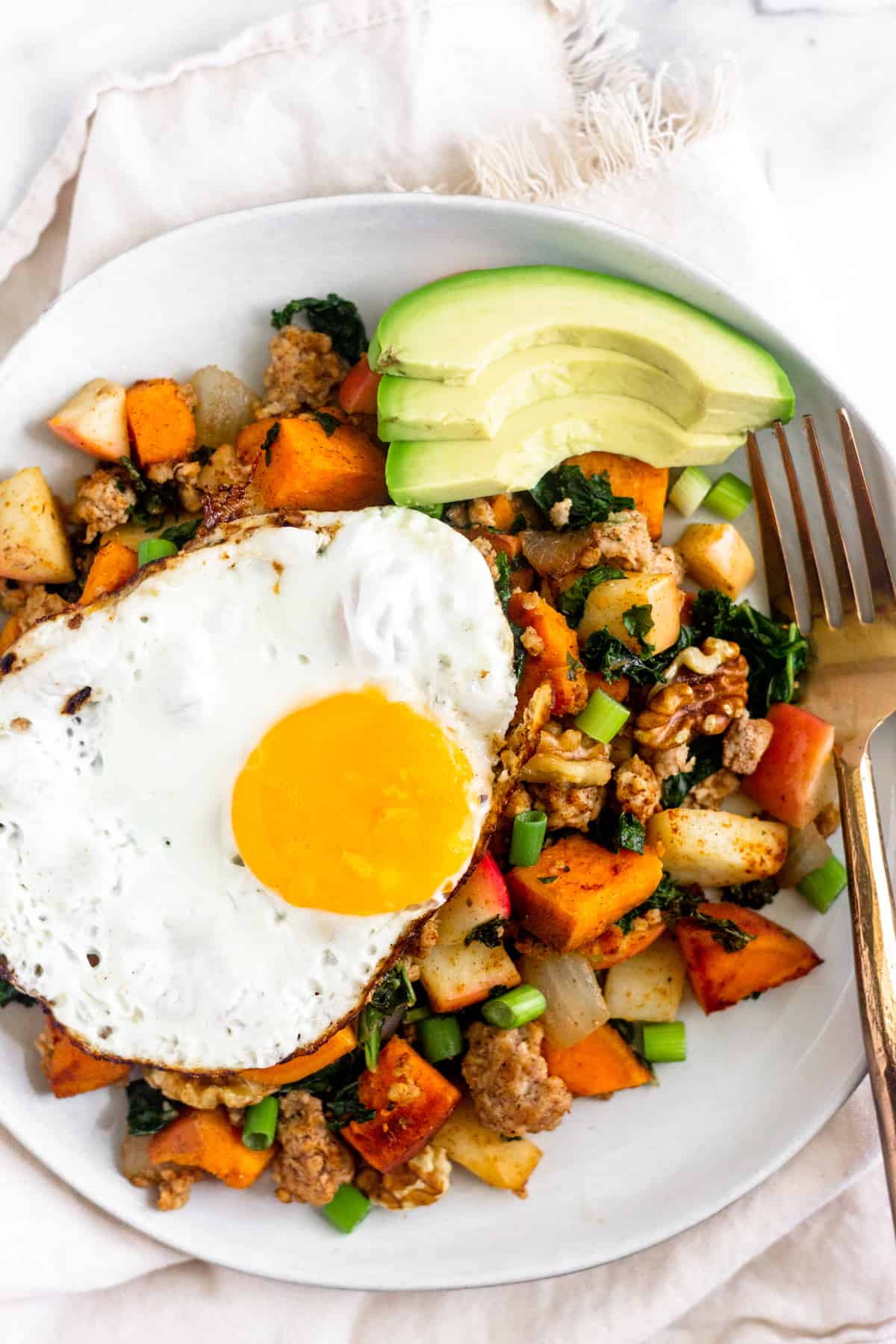 White plate on a tan linen filled with Whole30 Sweet Potato Sausage Hash, a fried egg, sliced avocado, and a bronze fork.