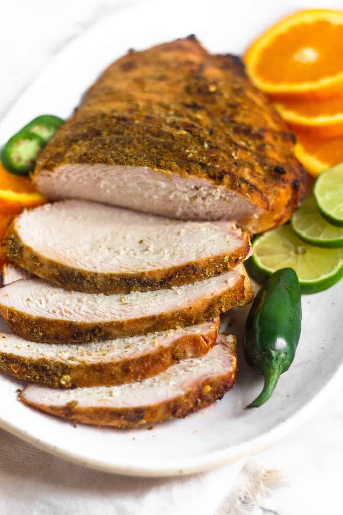 A platter of citrus grilled turkey breast with a few pieces cut laying on a plate next to some sliced limes, oranges, and a jalapeño.