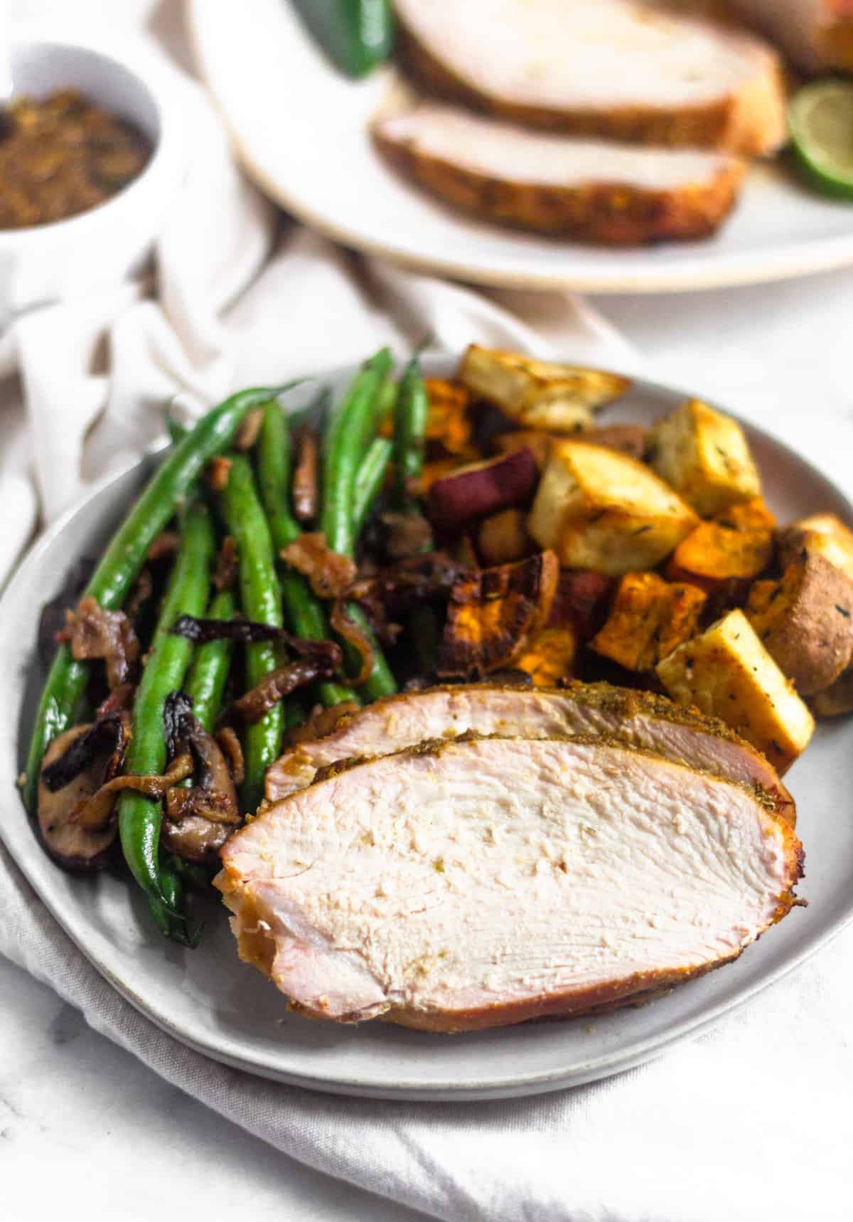 A plate of sliced citrus grilled turkey breast with a side of green beans and roasted sweet potatoes. Behind it is a platter os sliced turkey breast and a bowl of sauce