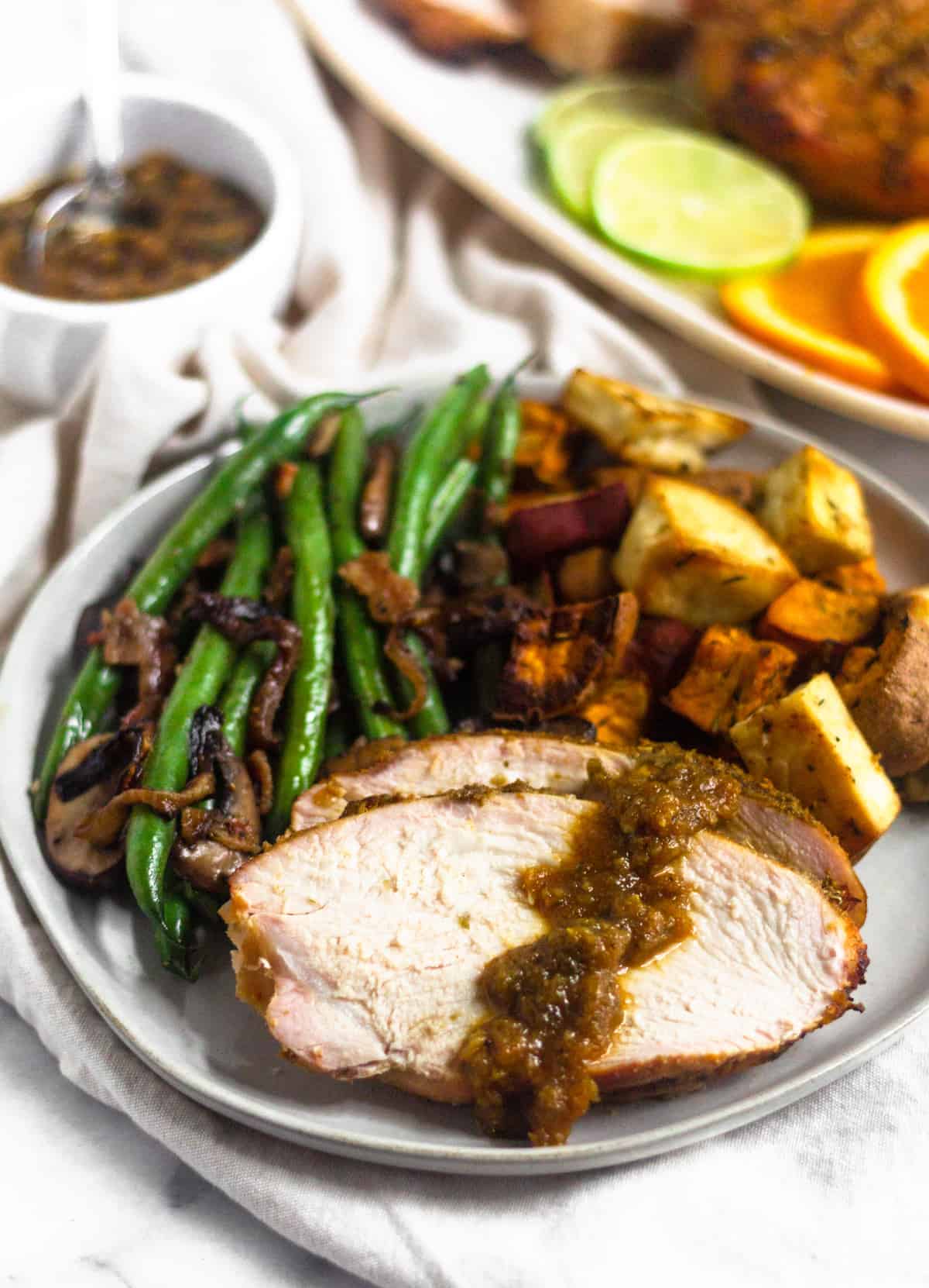 A plate of sliced citrus grilled turkey breast with a citrus sauce on it and a side of green beans and roasted sweet potatoes. Behind it is a platter os sliced turkey breast and a bowl of sauce