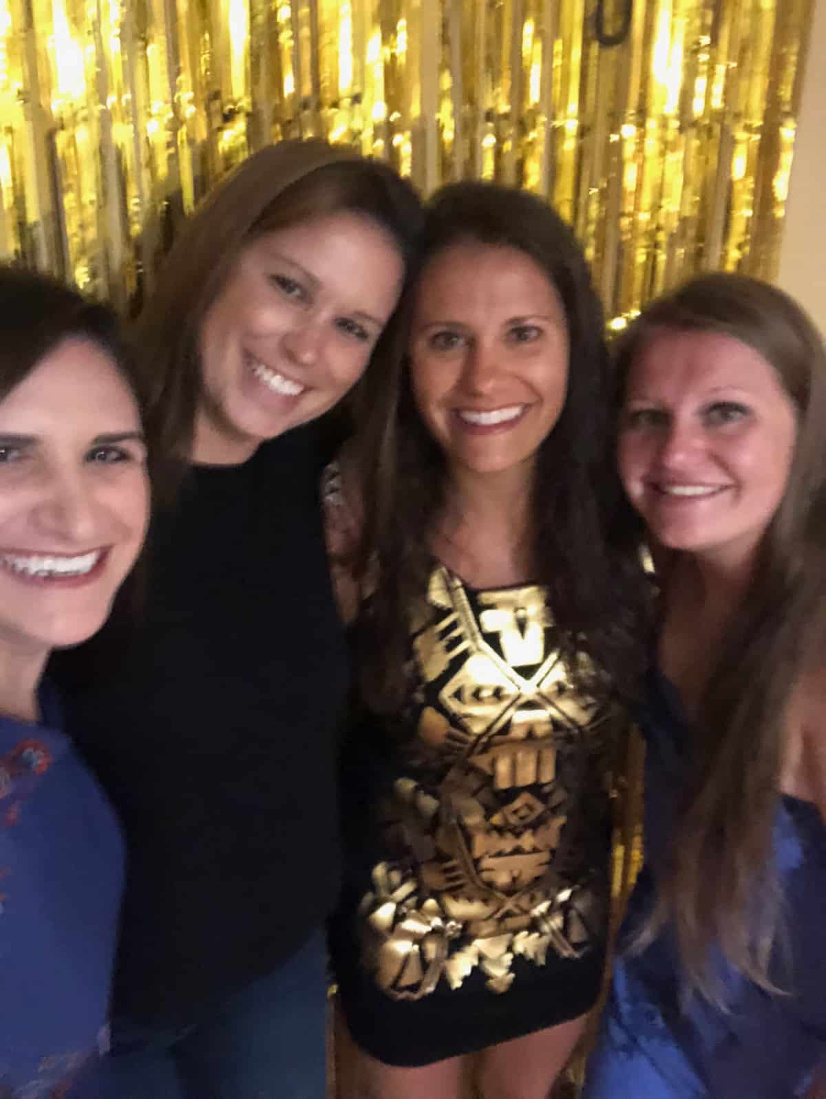 Four dark haired girls in front of a gold streamer, They are dressing up and ready to go out.