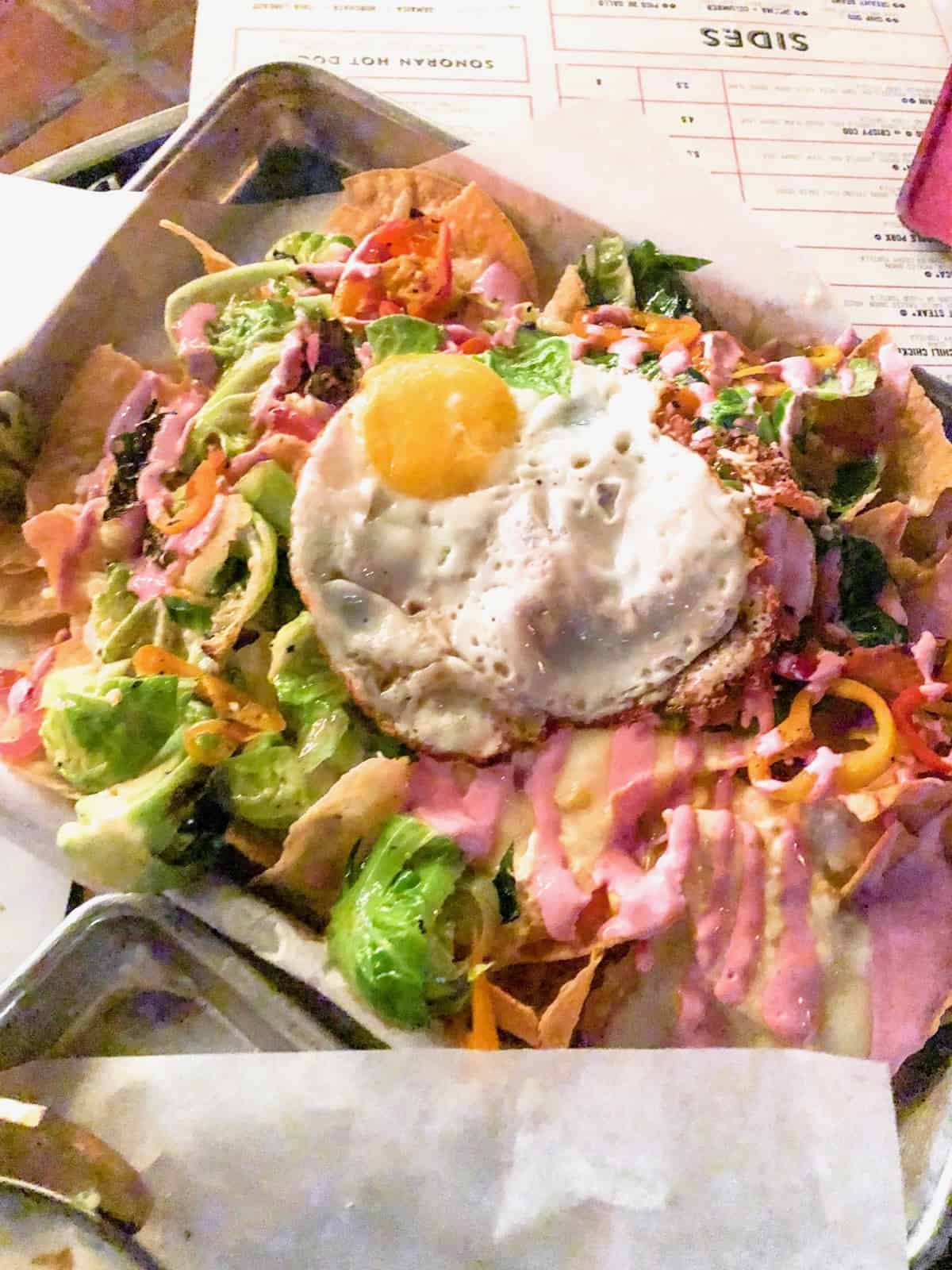 Plate of brussels sprouts nachos - chips with brussels sprouts, peppers, pickled onions, queso, beet crema, and a fried egg