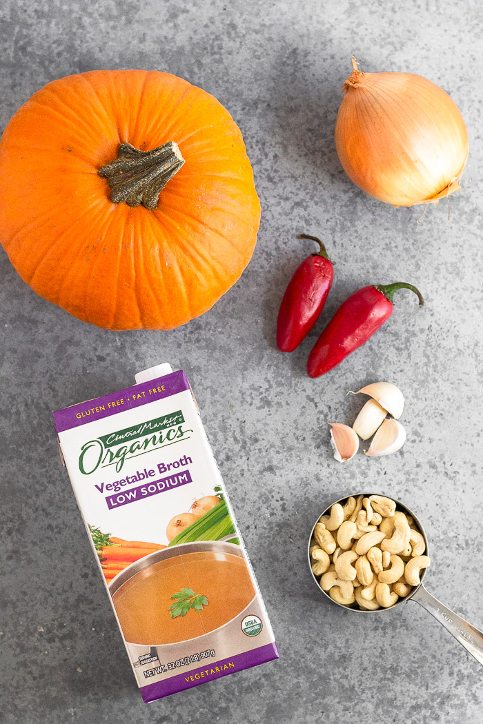Grey surface with a pumpkin, whole onion, two chili peppers, cup of cashews, garlic cloves, and a carton of broth.